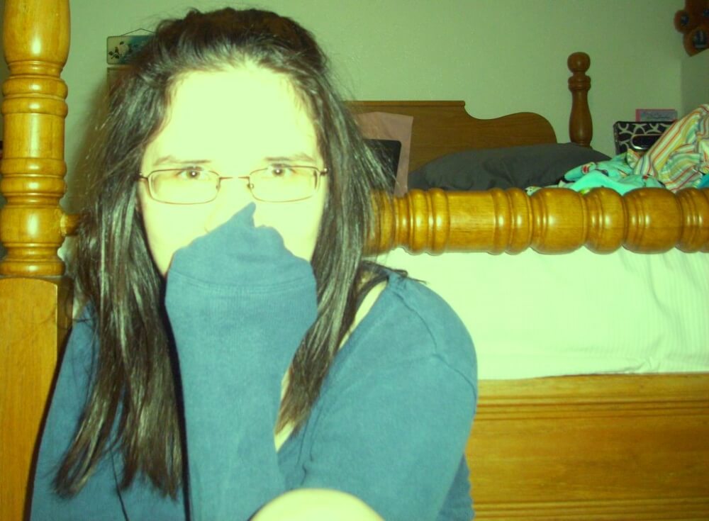 A photo of myself sitting at the foot of my bed. My hair's in a half-ponytail. I'm in a blue sweater; my mouth is covered by my first.