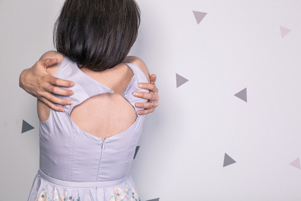 Person with black shoulder-length hair and light grey dress hugging self in front of white backdrop with grey triangles
