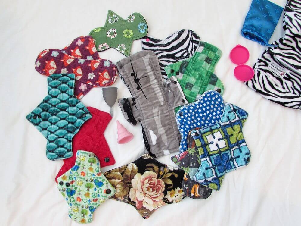 12 cloth pads, assorted, plus two menstrual cups laying on white surface in a circle collage