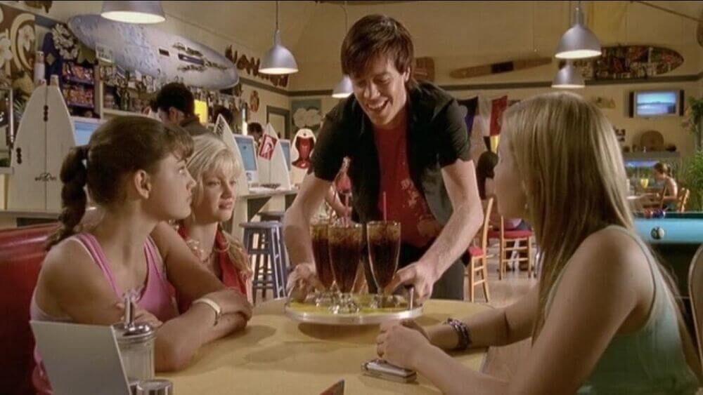 Zane placing a tray of three brown-colored drinks on the girls' table