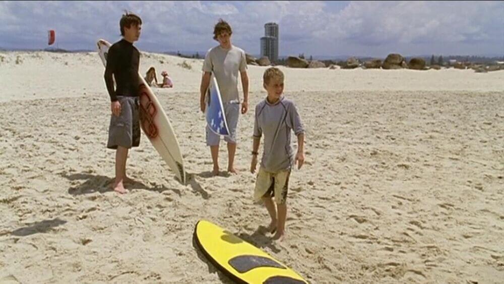 Zane and Nate standing behind Elliot, who just washed up on shore with his surfboard