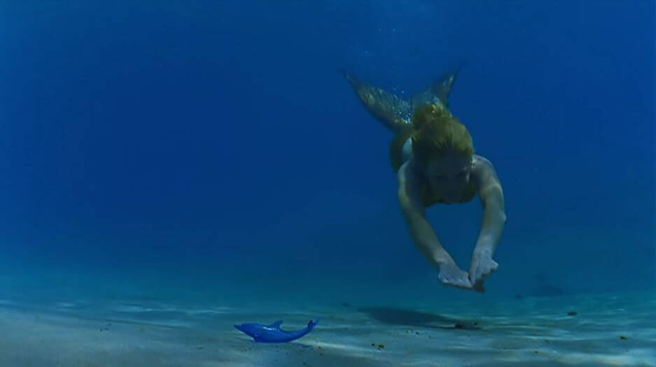 Rikki swimming, as a mermaid, underwater towards a small, blue dolphin figurine on the sand