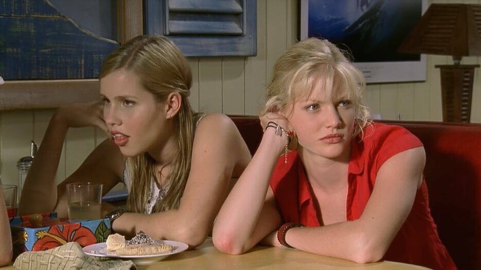 Emma and Rikki sit next to each other in a booth, Emma looking to Cleo and Rikki looking away from the table