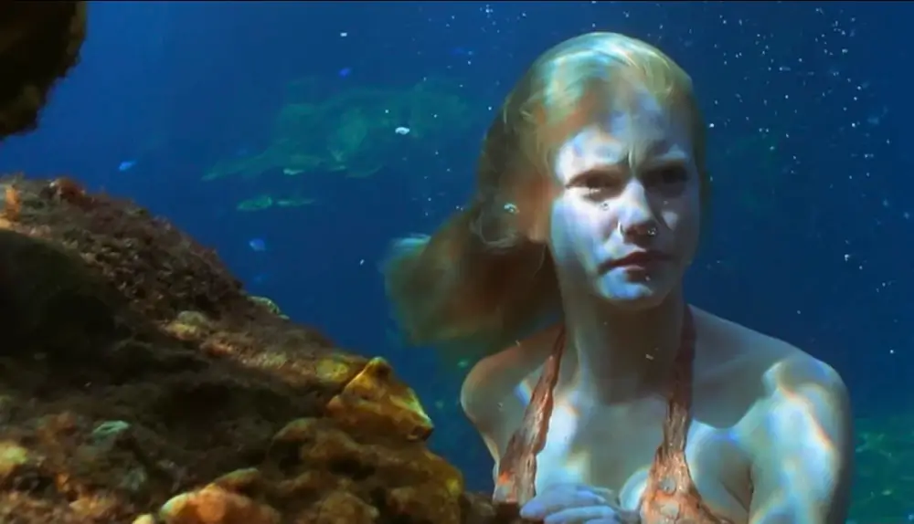 Rikki underwater, as a mermaid, slightly behind a reef