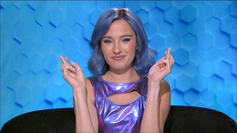 Cosette Rinab as Ainsley sitting on a couch in front of a blue wall with hexagonal textures, crossing fingers on both hands, wearing a blue shirt that shimmers, with shoulder-length blue hair